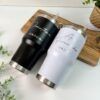 Dos vasos de acero inoxidable con tapa sobre una tabla de madera. Uno es negro y tiene la palabra novio y un nombre en letras blancas, el otro es blanco y tiene la palabra novia y un nombre en letras doradas. La escena está decorada con vegetación.