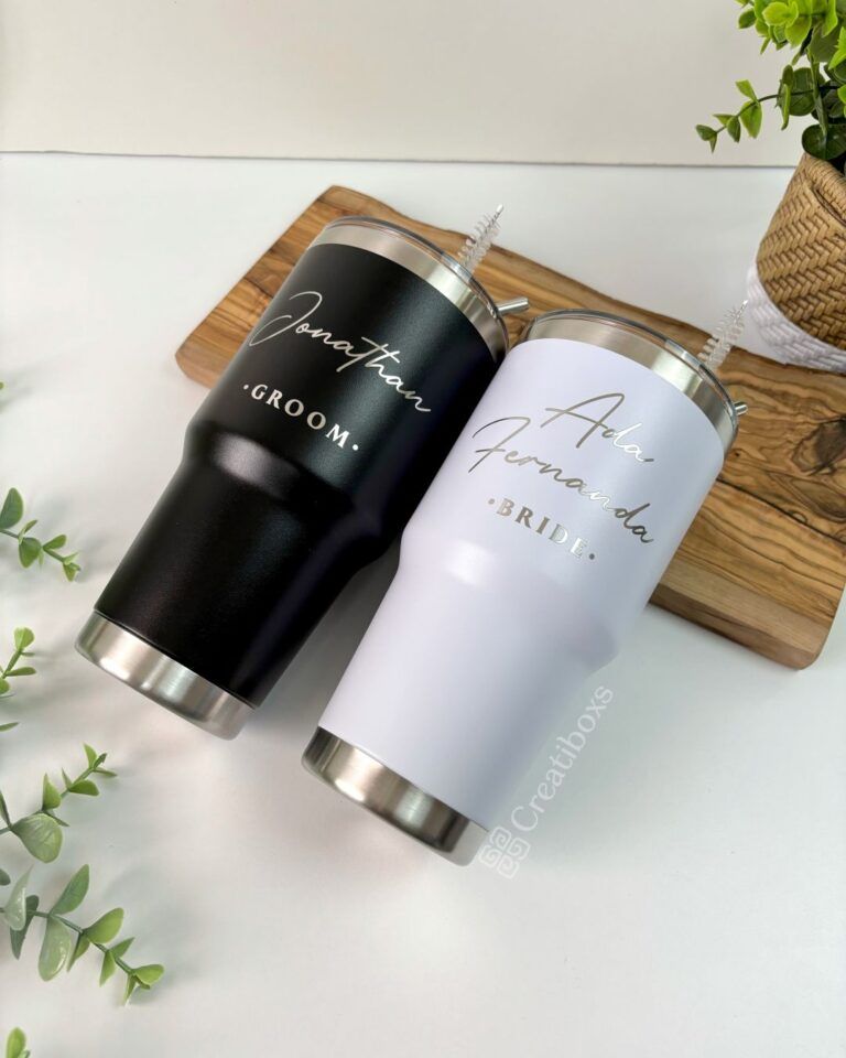Dos vasos de acero inoxidable con tapa sobre una tabla de madera. Uno es negro y tiene la palabra novio y un nombre en letras blancas, el otro es blanco y tiene la palabra novia y un nombre en letras doradas. La escena está decorada con vegetación.