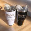Hay dos tazas de viaje sobre una superficie de madera. Una es blanca y dice Bride Adriana & Adrian y la otra es negra y dice Groom Adriana & Adrian. Ambas tienen pajitas de metal, tapas transparentes y la fecha es 20-10-2023.