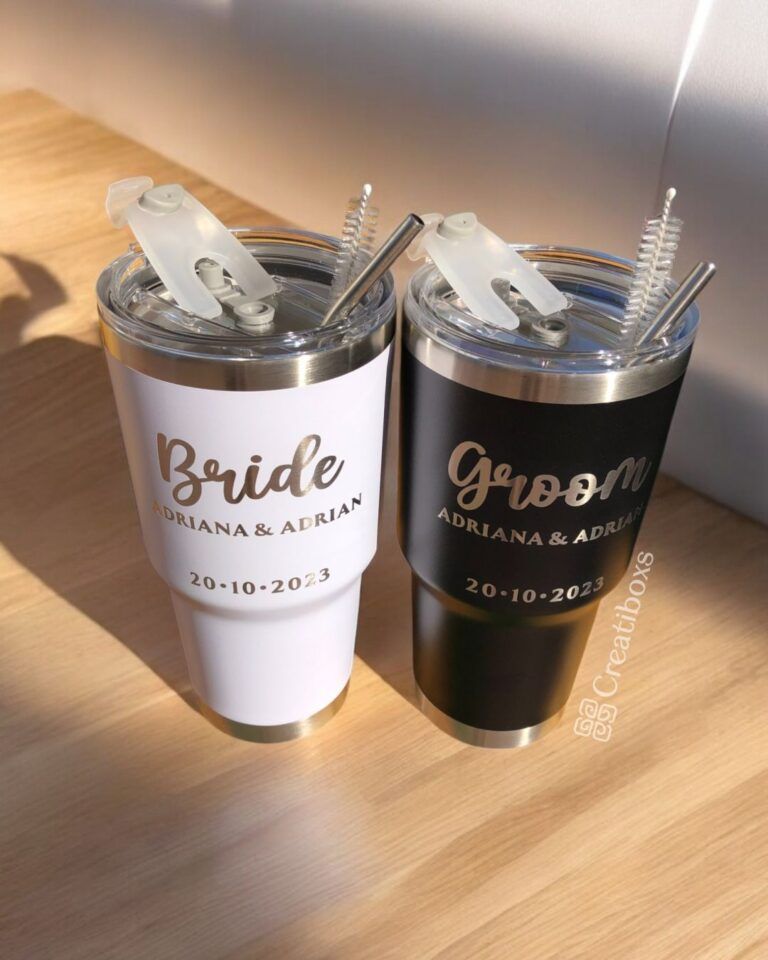 Hay dos tazas de viaje sobre una superficie de madera. Una es blanca y dice Bride Adriana & Adrian y la otra es negra y dice Groom Adriana & Adrian. Ambas tienen pajitas de metal, tapas transparentes y la fecha es 20-10-2023.