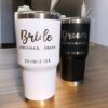 Dos vasos altos con aislamiento se encuentran sobre una superficie de madera. Uno es blanco con la inscripción Bride Adriana & Adrian 20-10-2023 en texto dorado, y el otro es negro con la inscripción Groom Adriana & Adrian 20-10-2023 en texto plateado. Ambos tienen pajitas de metal.