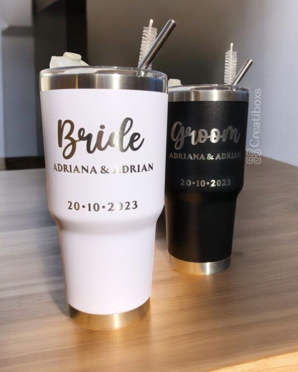 Dos vasos altos con aislamiento se encuentran sobre una superficie de madera. Uno es blanco con la inscripción Bride Adriana & Adrian 20-10-2023 en texto dorado, y el otro es negro con la inscripción Groom Adriana & Adrian 20-10-2023 en texto plateado. Ambos tienen pajitas de metal.