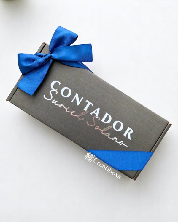 La caja de regalo rectangular de Tegalo corporativo con logo presenta un lazo de cinta azul y el logotipo “Contador Suriel Solano” elegantemente exhibido en la tapa.