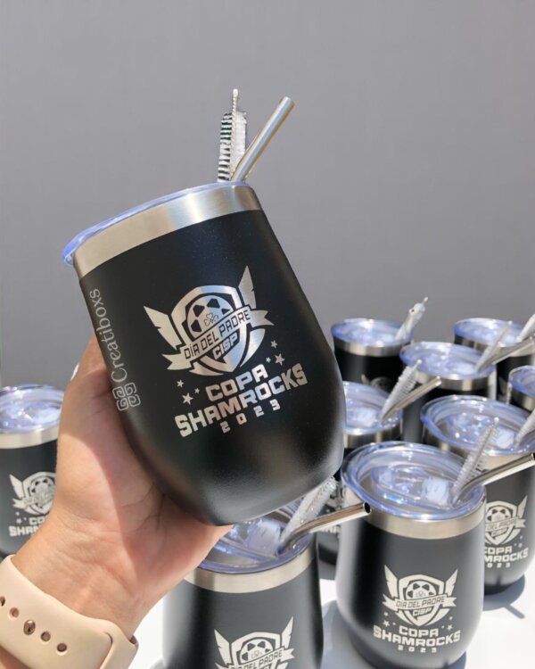 Una mano sostiene un vaso negro Copa Shamrocks 2023 "Termos personalizados para regalo corporativo con logo", con un logotipo destacado, exhibido entre otros termos personalizados sobre una mesa.