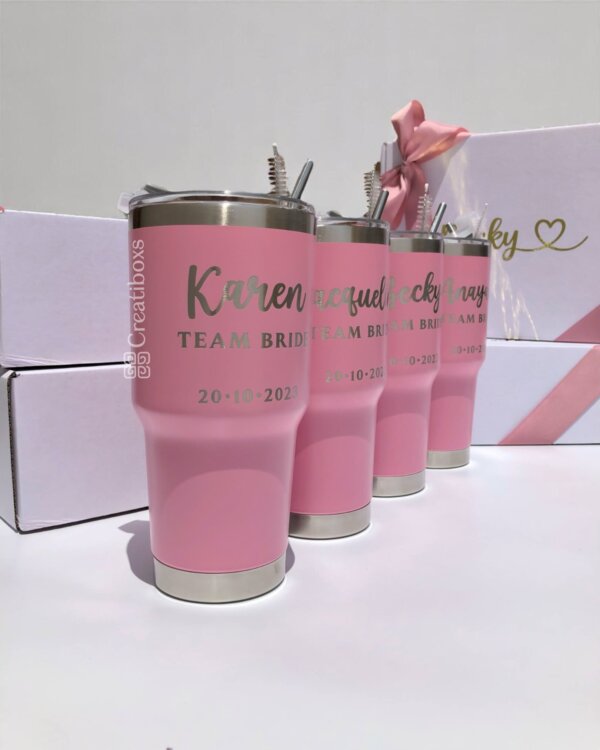 Cuatro termos rosados personalizados para boda con tapas metálicas, etiquetados TEAM BRIDE y una fecha, alineados en fila: recuerdos de boda ideales para su día especial.