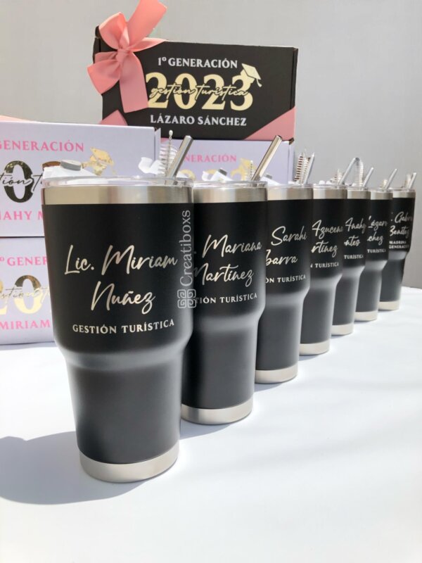 Seis Regalo de graduación termos personalizados en negro con nombres dorados, alineados frente a cajas de regalo con lazo rosa; un detalle elegante para graduaciones.