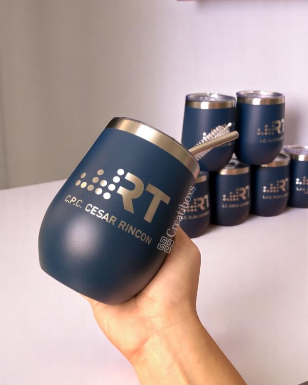Una mano sostiene uno de nuestros Termos personalizados para regalo corporativo con logo, que presenta un vaso azul con pajita; vasos personalizados similares se muestran en el fondo.