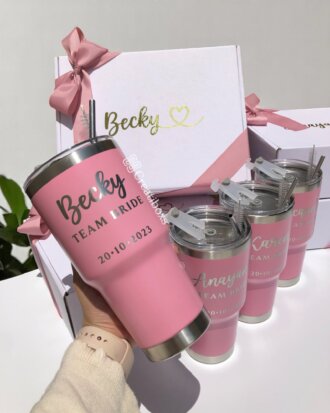 Cuatro Termos personalizados para boda color rosa con la frase Team Bride se exhiben frente a cajas de regalo decoradas con lazo rosa y el nombre Becky, ideales para obsequiar en celebraciones nupciales.