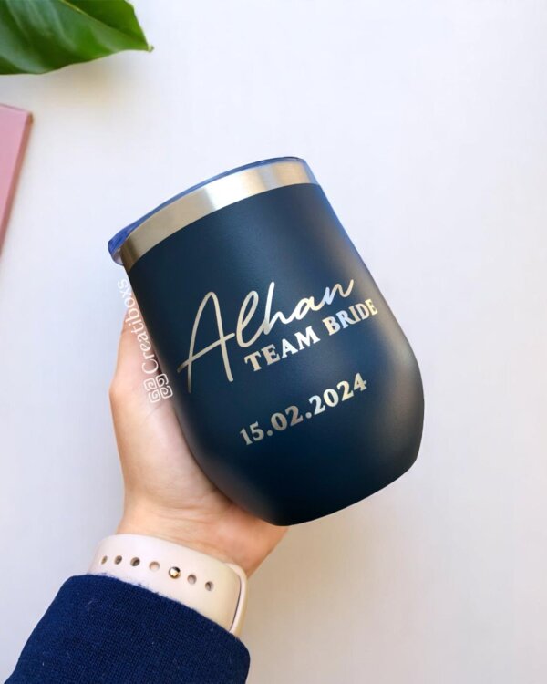 Una mano sostiene un vaso azul marino con la etiqueta "Alhan TEAM BRIDE 15.02.2024" en letra blanca, uno de nuestros termos personalizados para boda, ideal como un regalo memorable para celebraciones de bodas.