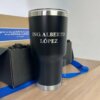 Un vaso negro del Kit termo clásico 590ml y caja chica personalizada, grabado con ING. ALBERTO LÓPEZ, descansa sobre un escritorio de madera con un lazo azul al lado.
