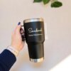 Mano sosteniendo el Regalo del día del padre con termo personalizado, con impreso "Sandoval Esposo | Padre | Birrero"; planta frondosa al fondo: regalo ideal para el Día del Padre.