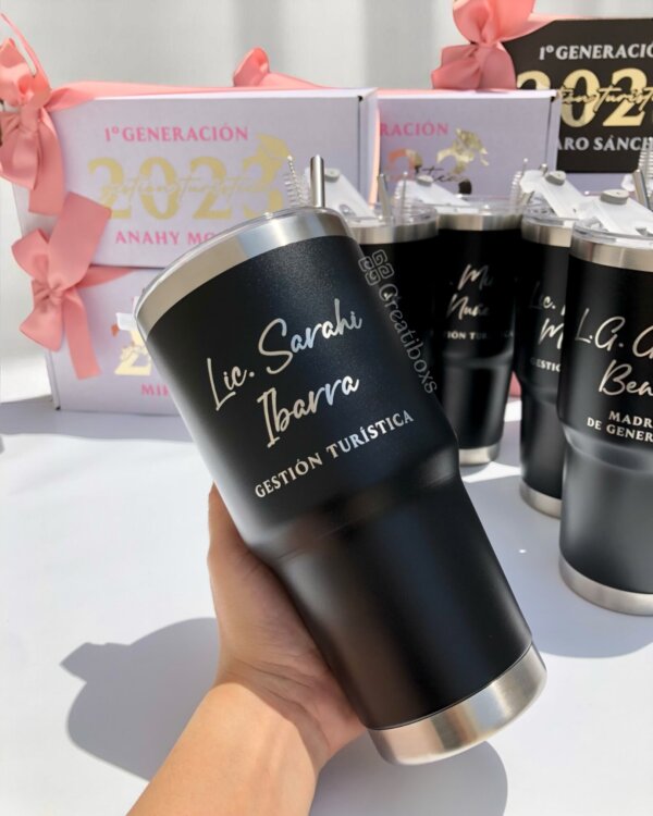 Una mano sostiene el “Regalo de graduación termos personalizados” (un vaso negro con texto dorado); en el fondo, cajas de regalo con lazos rosas lo resaltan como un regalo de graduación ideal.