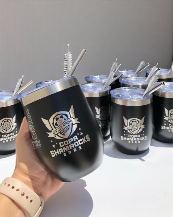 Una mano sostiene un termo personalizado con logo negro "Copa Shamrocks 2023" entre varios termos personalizados para regalo corporativo con logo idénticos.
