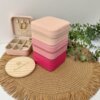 Estuches Joyero personalizado de cuero sintético rosa apilados sobre un tapete tejido, con vegetación y un estuche abierto con pendientes de oro.