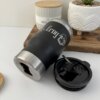 Un "Termo personalizado portalatas 4 En 1 con destapador 420ml 14oz" negro con "Erny" impreso en él se sienta al lado de su tapa desprendida en una superficie blanca.
