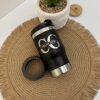 Termo negro personalizado portalatas 4 en 1 con destapador (420ml/14oz) con el logotipo de Carlos Gómez, situado sobre una alfombrilla tejida junto a una maceta y un posavasos, que ofrece un estilo versátil y funcional para sus bebidas.