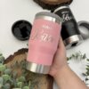 Una mano sostiene un vaso rosa del Kit termo portalatas x2 y caja personalizada chica magnética; cerca hay un vaso negro y plantas sobre superficies de madera.