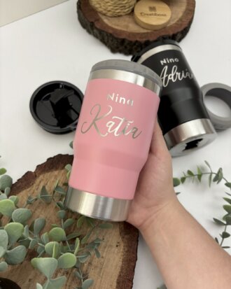 Una mano sostiene un vaso rosa del Kit termo portalatas x2 y caja personalizada chica magnética; cerca hay un vaso negro y plantas sobre superficies de madera.