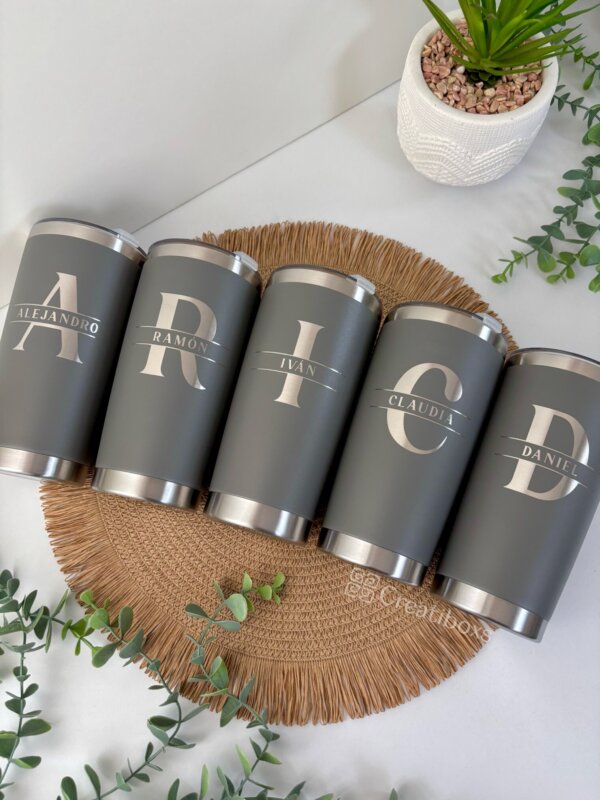 Cinco termos personalizados para boda en color gris, personalizados con nombres e iniciales, ideales como regalos de boda, dispuestos sobre un tapete de mimbre con plantas verdes cerca.