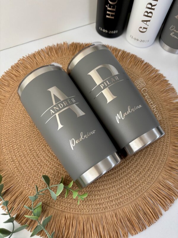 Dos Termos personalizados para boda en gris, etiquetados Andrés Padrino y Pilar Madrina, descansan sobre un mantel individual tejido, un recuerdo ideal para cualquier celebración de boda.
