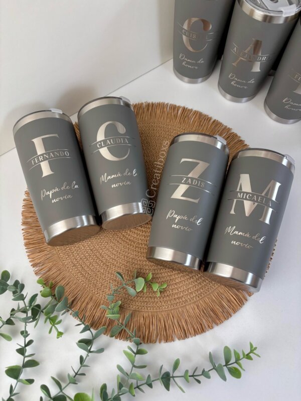 Cuatro termos personalizados para boda sobre un tapete tejido con vegetación, etiquetados para los padres de los novios: recuerdos de boda únicos que añaden un toque especial a cualquier celebración.