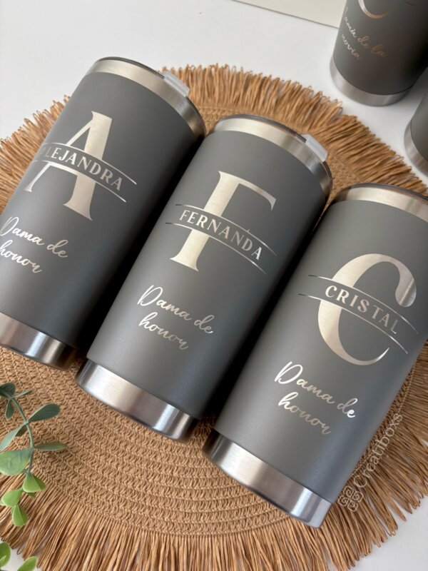 Tres termos personalizados para boda en color gris, cada uno con un nombre y una "Dama de honor", están dispuestos sobre un tapete tejido, perfecto para cualquier celebración de boda.
