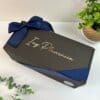 Una caja de regalo negra con el nombre Ing. Plasencia escrito en la tapa en una elegante letra, atada con una cinta azul oscuro, se coloca sobre una superficie blanca al lado de plantas verdes en macetas y ramitas de vegetación.