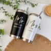 Celebra tu boda con los Termos personalizados para boda: un termo “Mr.” negro y otro “Mrs.” blanco, ambos con nombres y fecha, presentados sobre un tapete tejido.