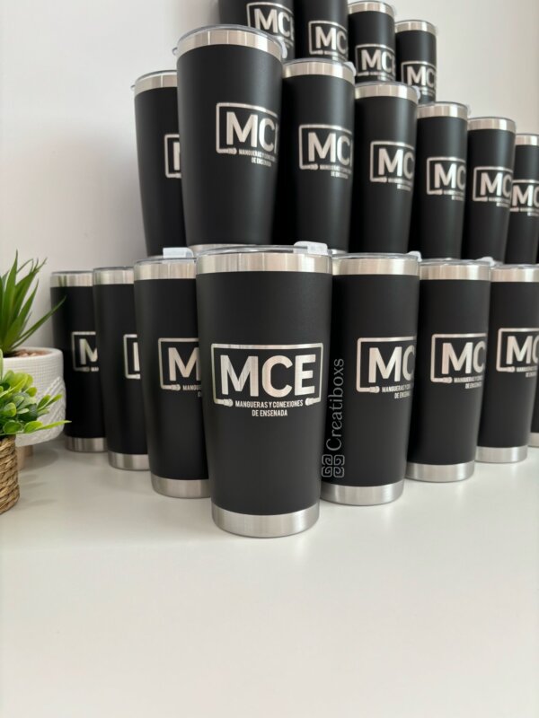 Termos personalizados para regalo corporativo con logo “MCE” apilados sobre una superficie blanca, junto a una pequeña planta.