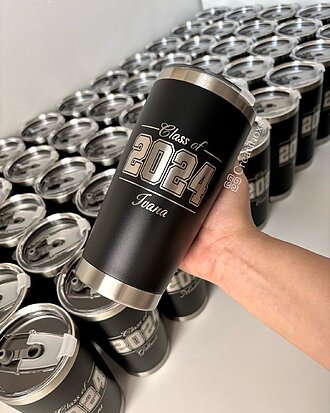 Una mano sostiene uno de los Termos personalizados de graduación regalo de graduación en color negro con el nombre "Class of 2024 Tania", rodeado de termos idénticos, perfecto para regalar en celebraciones de fin de curso.