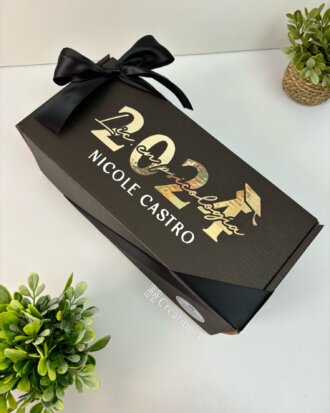 Regalo de graduación con caja negra con “2024” y nombre en dorado, decorado con listón negro y plantas. Ideal para un recuerdo memorable en tu celebración especial.
