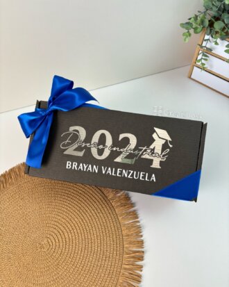 Regalo de graduación con caja negra con moño azul, 2024 y nombre en la tapa, sobre mantel tejido.