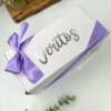 Una caja de regalo blanca con Veritos en la parte superior y una cinta violeta, rodeada de plantas sobre una superficie blanca, constituye el regalo perfecto de Kit termo clásico grande 890ml y caja chica personalizada.