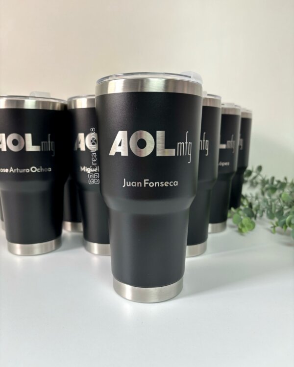 Termo personalizado para regalo corporativo con logo, modelo negro metálico rotulado "AOL mfg Juan Fonseca", rodeado de otros termos similares, ideal como obsequio empresarial sobre superficie blanca.