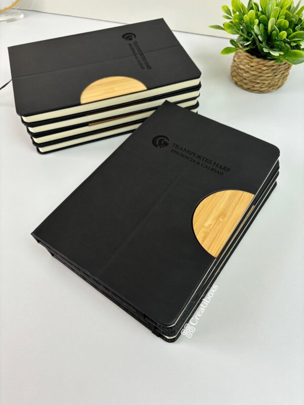 Libretas personalizadas apiladas para regalo corporativo con logotipo, con cubiertas negras con detalles de madera y una planta verde en maceta sobre un escritorio blanco, perfectas para cualquier espacio de trabajo profesional.