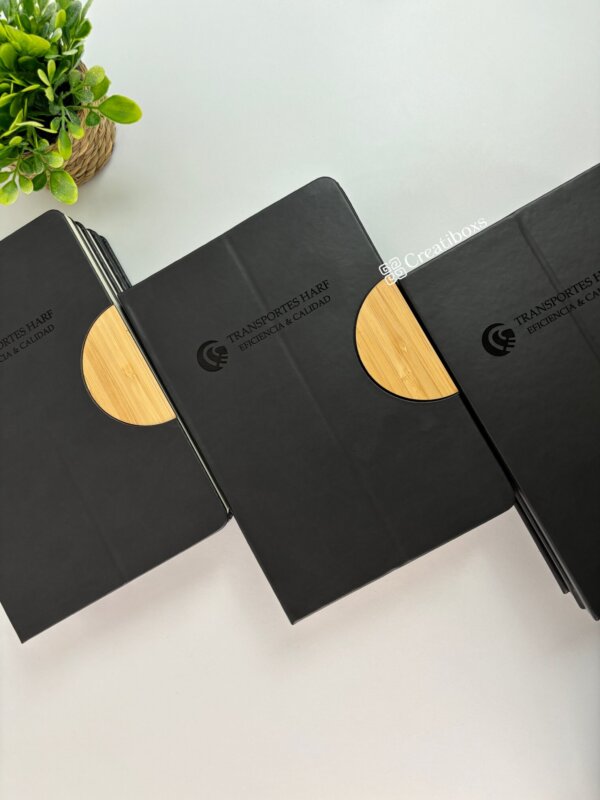 Libretas personalizadas para regalo corporativo con logo y detalles de madera en negro, exhibidas junto a una planta sobre fondo blanco