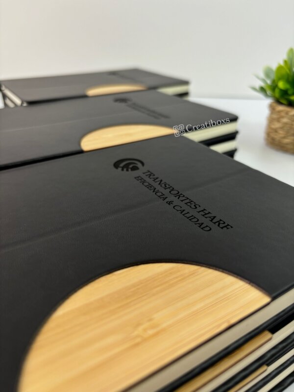 Libretas personalizadas para regalo corporativo con logo, en negro y textura madera, reposan sobre un escritorio junto a una planta—ideales para destacar tu marca o como obsequio empresarial.
