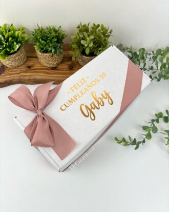 Presenta caja personalizada en una elegante caja blanca con listón rosa, texto dorado con frase de feliz cumpleaños y pequeñas plantas de fondo: el detalle ideal para regalar.