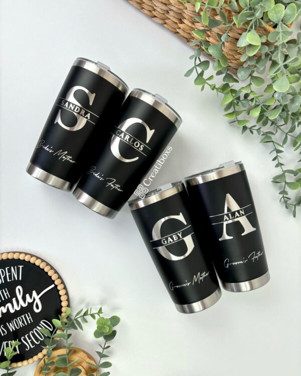 Cuatro termos personalizados para boda en color negro, personalizados con nombres e iniciales, están dispuestos sobre una superficie blanca entre plantas verdes, perfectos como regalos de boda únicos.