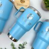 Tres termos personalizados para regalo corporativo con logo TRANSPORTES HAFE y tapa azul, sobre superficie blanca con plantas verdes—ideales como obsequio empresarial.