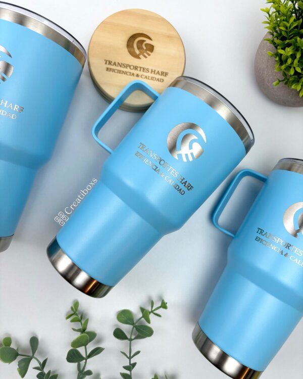 Tres termos personalizados para regalo corporativo con logo TRANSPORTES HAFE y tapa azul, sobre superficie blanca con plantas verdes—ideales como obsequio empresarial.