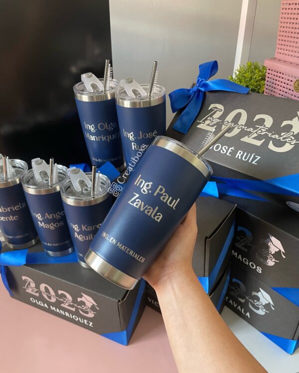 Una mano sostiene un "Regalo de graduación termos personalizados" azul grabado con el Ing. Paul Zavala, con más vasos personalizados perfectos para regalos de graduación visibles en el fondo.