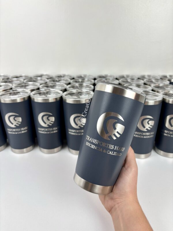 Una mano sostiene un Termo personalizados para regalo corporativo con logo azul marino, rodeado de otros sobre una superficie blanca, ideal como regalos empresariales.