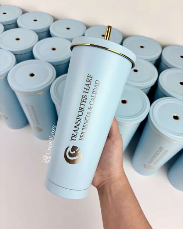 Una mano sostiene un termo personalizado para regalo corporativo con logo en color azul claro y popote dorado, rodeado de termos similares. Texto: TRANSPORTES HARF—ideal para regalos empresariales.