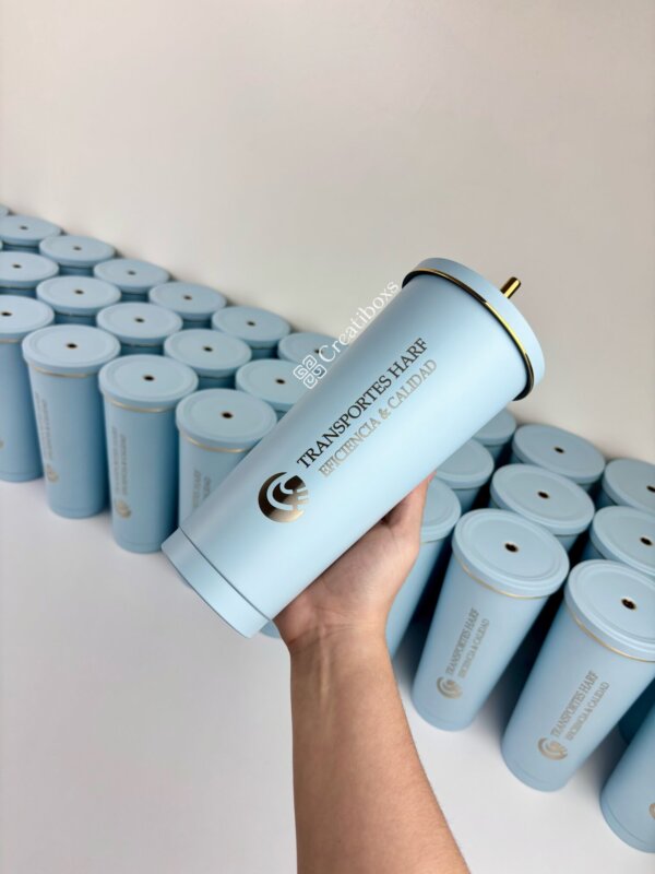 Una mano sostiene uno de los termos personalizados para regalo corporativo con logo, de color azul, entre varios similares alineados sobre una superficie blanca.
