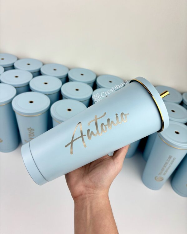 Una mano sostiene uno de nuestros termos personalizados para regalo corporativo con logo, en color azul claro y rotulado “Antonio”; ideal para empresas, rodeado de otros vasos similares.
