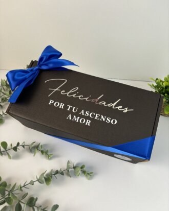 Celebra con el Kit Termo Clásico Grande de 890 ml y caja chica personalizada: una caja negra con un lazo azul y la frase "Felicidades por tu ascenso, amor", rodeada de vegetación. Ideal para ocasiones especiales y memorables.