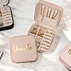 Un joyero rosa con la etiqueta Danielle contiene anillos, aretes y collares, organizados en compartimentos separados. Se encuentra sobre una superficie de mármol junto a otros joyeros.
