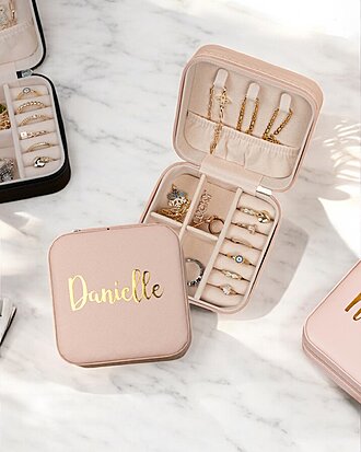 Un joyero rosa con la etiqueta Danielle contiene anillos, aretes y collares, organizados en compartimentos separados. Se encuentra sobre una superficie de mármol junto a otros joyeros.