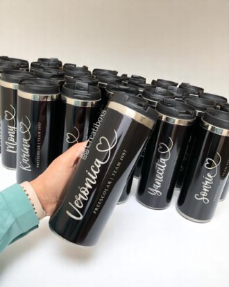 Termos personalizados para Reunión de generación: Una mano sostiene un termo negro personalizado con nombre y texto Preescolar Team 1997, en el fondo hay dispuestos más termos similares.